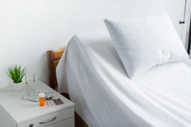 Pills and glass of water on bedside table near bed in hospital ward  clipart