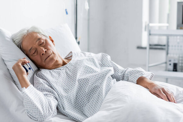 Пожилой азиатский пациент с пульсоксиметром спит на кровати в больнице 