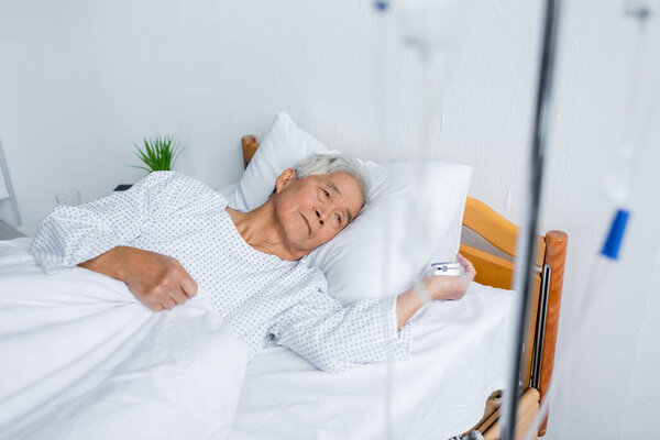 Старший азиатский мужчина с пульсоксиметром, лежащий рядом с размытой внутривенной терапией на кровати в больнице 