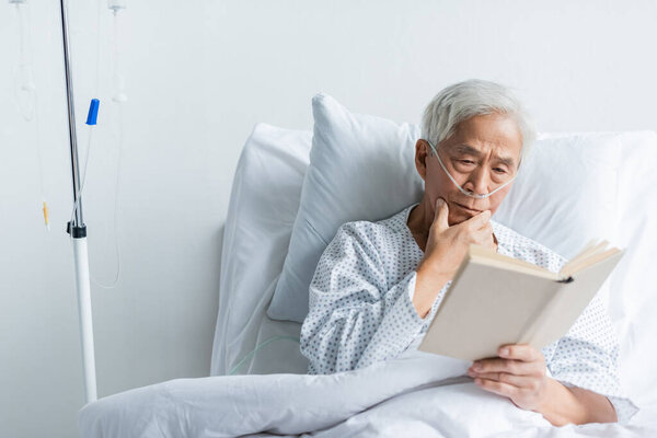 Пожилой азиатский пациент с носовой канюлей читает книгу на кровати в клинике 