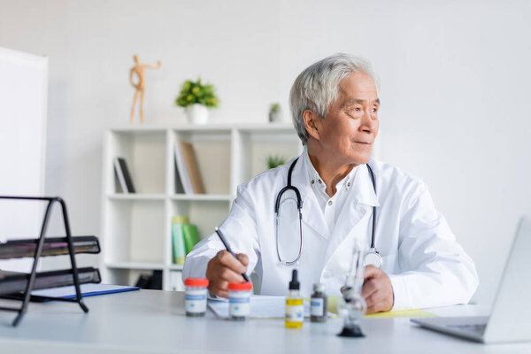 Asian doctor holding pen near cbd oil, bong and laptop in clinic 
