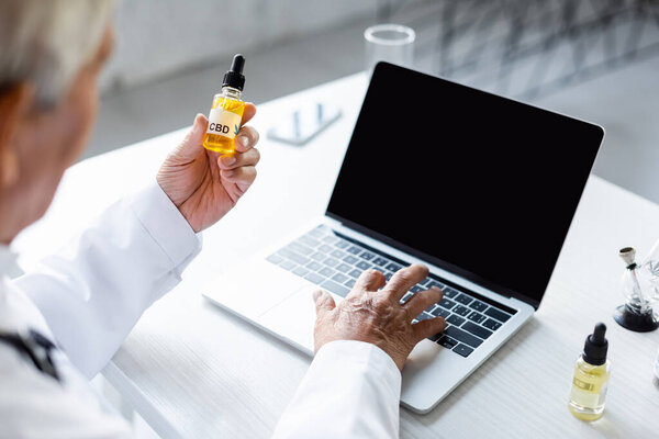 Blurred doctor holding cbd oil and using laptop with blank screen in clinic 