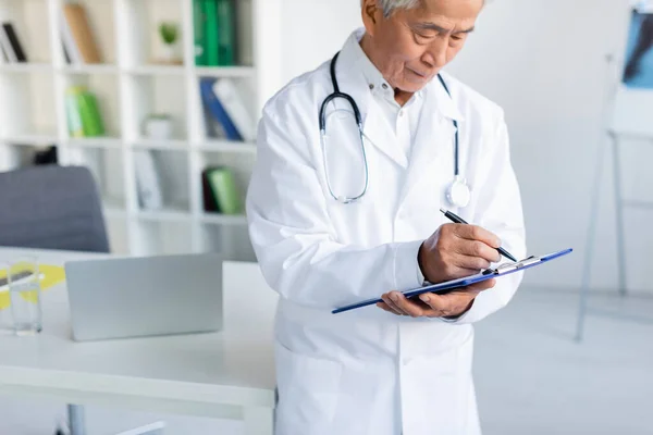 Elderly Asian Doctor Writing Clipboard Blurred Laptop Clinic — Stock Photo, Image