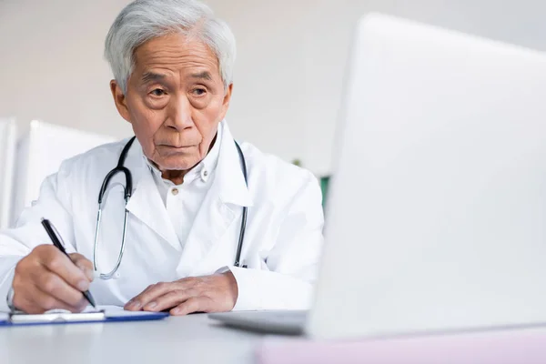 Anziani Asiatico Medico Writing Clipboard Blurred Laptop Hospital — Foto Stock