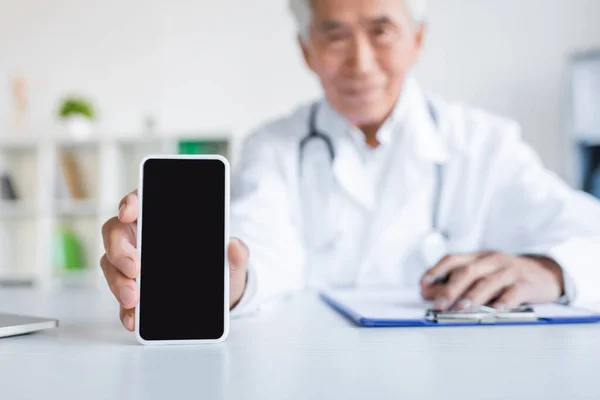 病院でクリップボードの近くに空白の画面とスマートフォンを保持しているアジアの医師のぼやけた — ストック写真