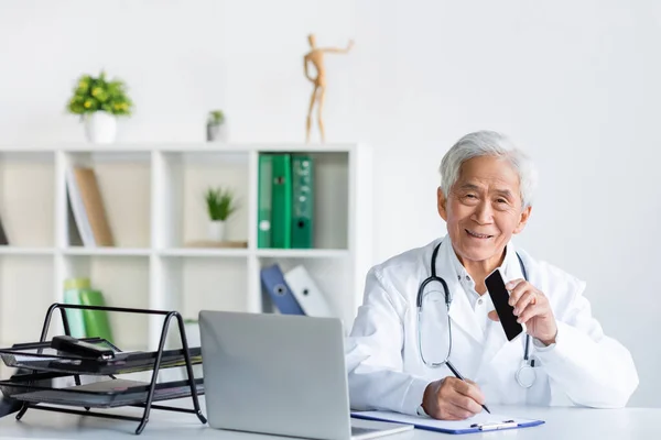 Smiling Asian Doctor Holding Smartphone Blank Screen While Writing Clipboard — Stock Photo, Image