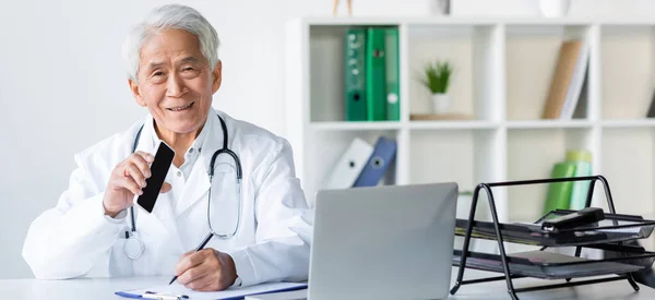 Smiling Asian Doctor Holding Smartphone While Writing Clipboard Laptop Hospital — Stock Photo, Image