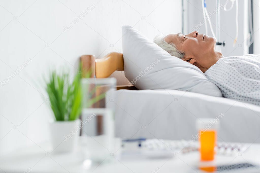 Side view of senior asian patient sleeping on bed near blurred water and pills in clinic 