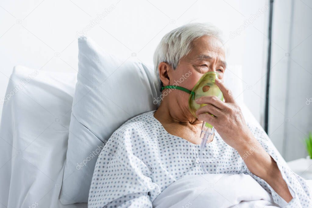 Senior asian patient holding oxygen mask while sitting on bed in hospital 