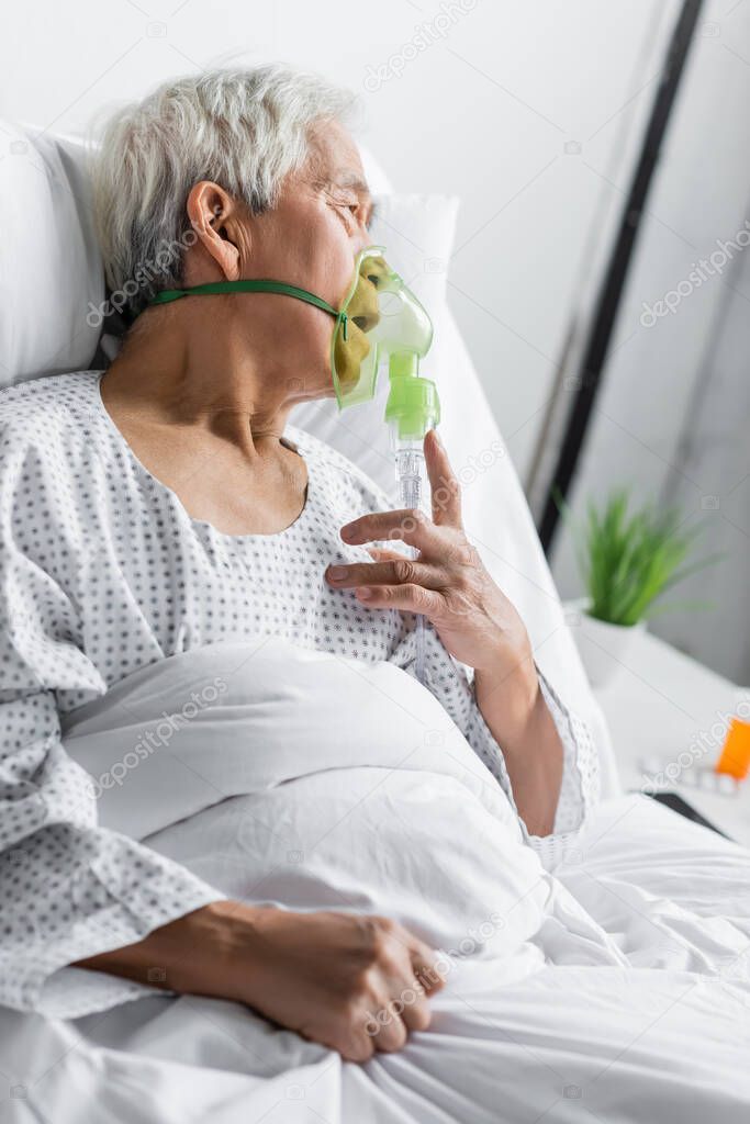 Side view of asian patient in oxygen mask lying on bed in hospital ward 