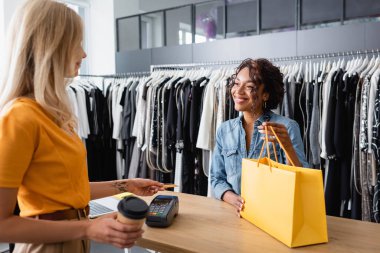 Dövmeli ve sarışın müşteri kredi kartıyla ödeme yapıyor. Butikte gülümseyen Afro-Amerikan satış asistanının yanında.