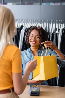 Gülümseyen Afrikalı Amerikalı satıcı butikte sarışın müşteriye sarı kağıt çanta veriyor.