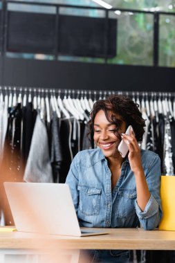 Neşeli Afrikalı Amerikalı satış elemanı akıllı telefondan konuşuyor ve giysi mağazasında dizüstü bilgisayar kullanıyor.