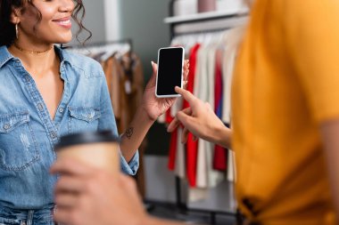 Akıllı telefonu işaret eden, neşeli Afro-Amerikan dükkan asistanının yanında boş ekranlı bir kadın görüntüsü. 