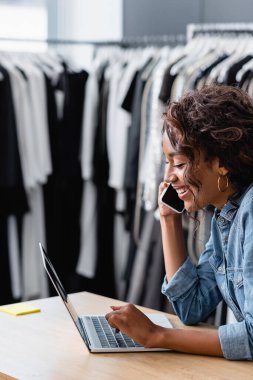 Mutlu Afrikalı Amerikalı kadın akıllı telefonla konuşuyor ve satış masasında dizüstü bilgisayar kullanıyor.