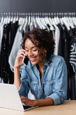 Neşeli Afrikalı Amerikalı kadın akıllı telefonla konuşuyor ve satış masasında dizüstü bilgisayar kullanıyor.