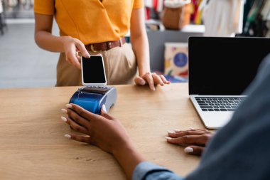 Bilgisayarın yanında akıllı telefondan ödeme yapan kadın ve butikte çalışan Afro-Amerikan satış elemanı.
