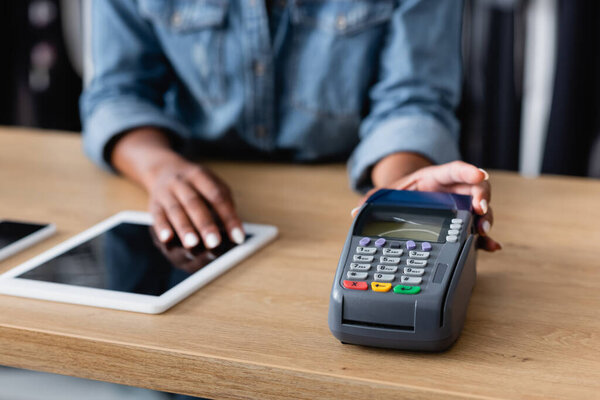 cropped view of african american saleswoman holding payment terminal near gadgets on sales counter desk