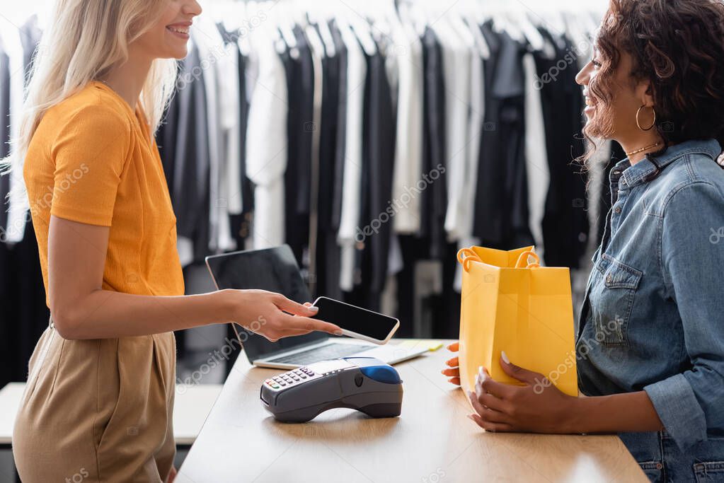 happy blonde woman paying with smartphone near african american saleswoman in boutique