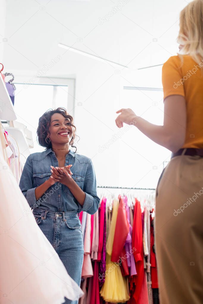 happy african american shop assistant with clenched hands looking at blurred customer gesturing in boutique 