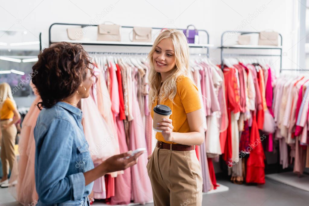 blonde woman holding paper cup and looking at blurred african american shop assistant with smartphone 