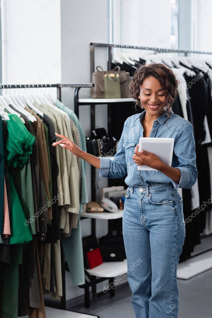 cheerful african american saleswoman holding digital tablet near clothing on rack