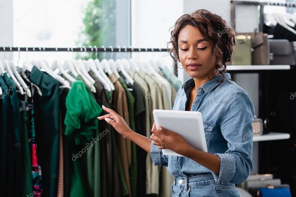 young african american saleswoman holding digital tablet and pointing at clothing on rack
