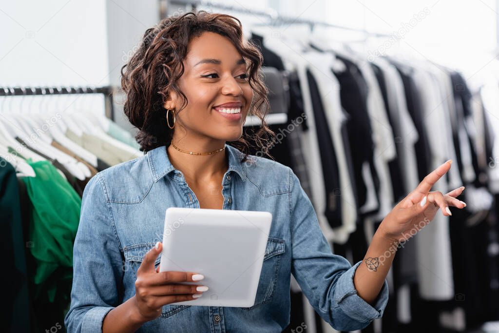 cheerful african american saleswoman with tattoo holding digital tablet near clothing on rack