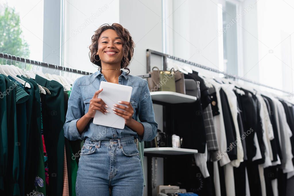 positive african american saleswoman holding digital tablet near clothing on rack