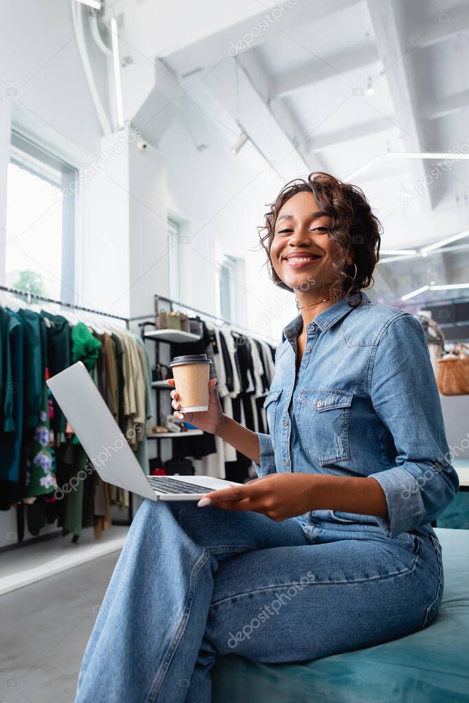 happy african american woman holding laptop and paper cup in clothing boutique 