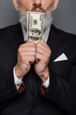 partial view of tattooed middle aged businessman kissing dollar banknotes isolated on grey clipart