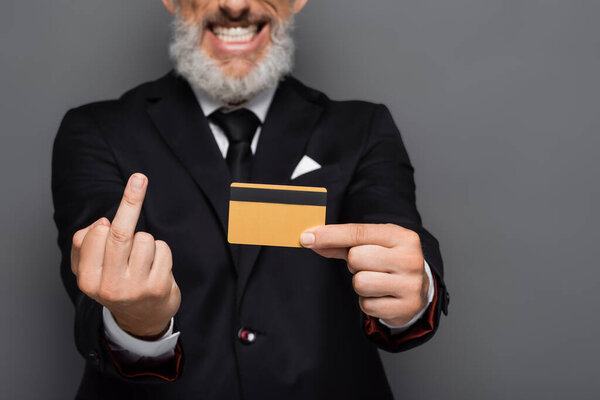cropped view of cheerful middle aged businessman in suit holding credit card while showing middle finger isolated on grey