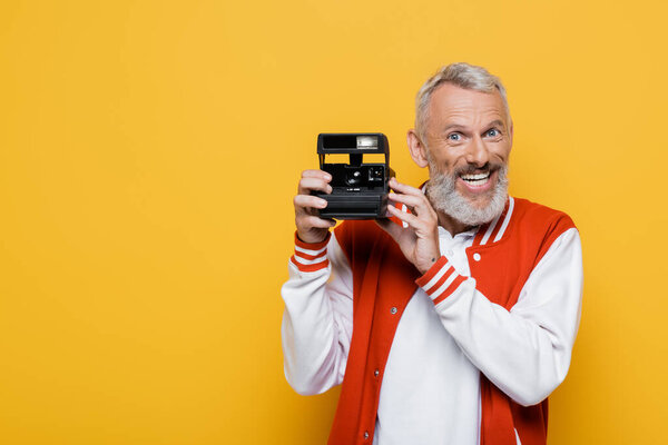 счастливый мужчина средних лет в пиджаке бомбардировщика с черной винтажной камерой, изолированной на желтом