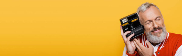 довольный мужчина средних лет в куртке бомбардировщика с черной винтажной камерой изолированы на желтый, баннер