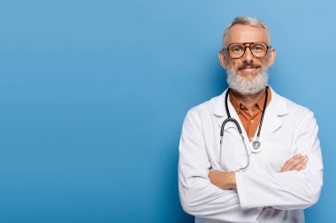 Beyaz önlüklü neşeli orta yaşlı doktor mavi kollarıyla ayakta duruyor.