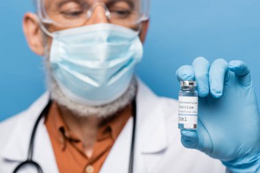 Tıbbi maskeli bulanık doktor ve lateks eldivenli şişe içinde koronavirüs aşısı olan mavi harfler.