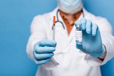 Doktorun lateks eldivenli, elinde Coronavirus aşısı şişesi ve mavi şırınga tutan görüntüsü.