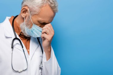 Üzgün orta yaşlı doktor beyaz önlüklü ve mavi maskeli.