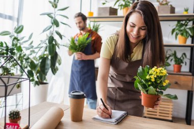 Young seller holding plant and writing on notebook near blurred colleague in flower shop  clipart
