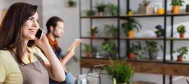 Smiling florist in apron talking on smartphone in blurred flower shop, banner  clipart