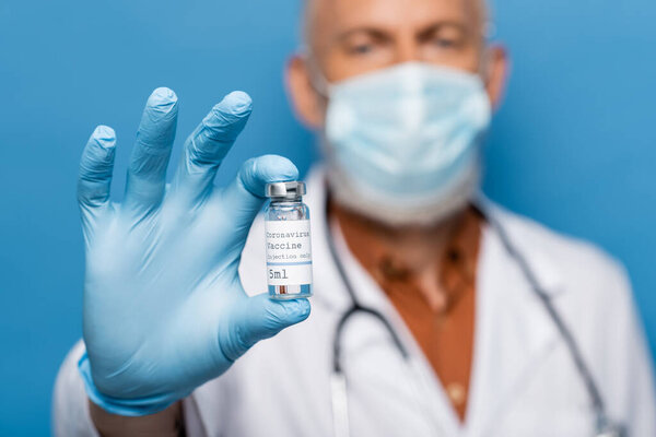 blurred doctor in medical mask holding vial with coronavirus vaccine isolated on blue