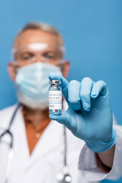 blurred doctor in medical mask and latex glove holding vial with coronavirus vaccine isolated on blue