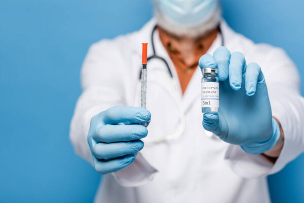 cropped view of doctor in latex gloves holding vial with coronavirus vaccine and syringe isolated on blue