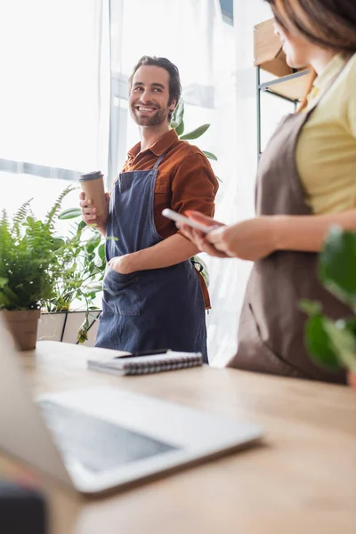 Smiling Retailer Paper Cup Looking Colleague Smartphone Flower Shop — Stock Photo, Image