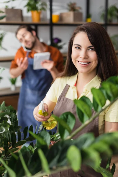 Florista Sonriente Delantal Sosteniendo Pulverizador Cerca Las Plantas Mirando Cámara —  Fotos de Stock