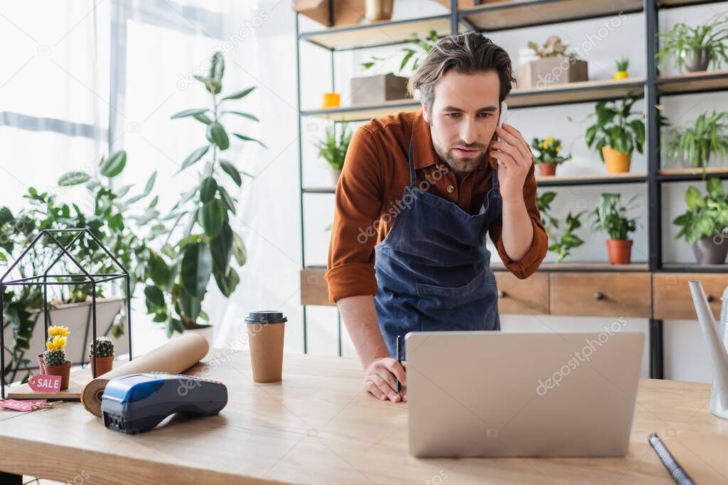 Seller in apron talking on smartphone near laptop and coffee in flower shop 
