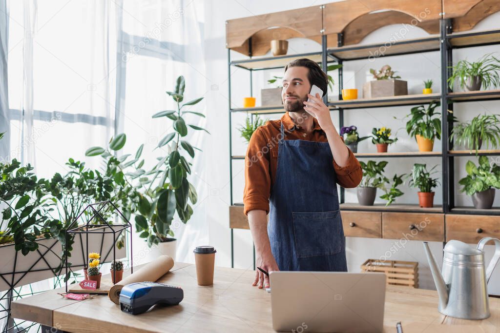 Seller in apron talking on smartphone near coffee and laptop in flower shop 