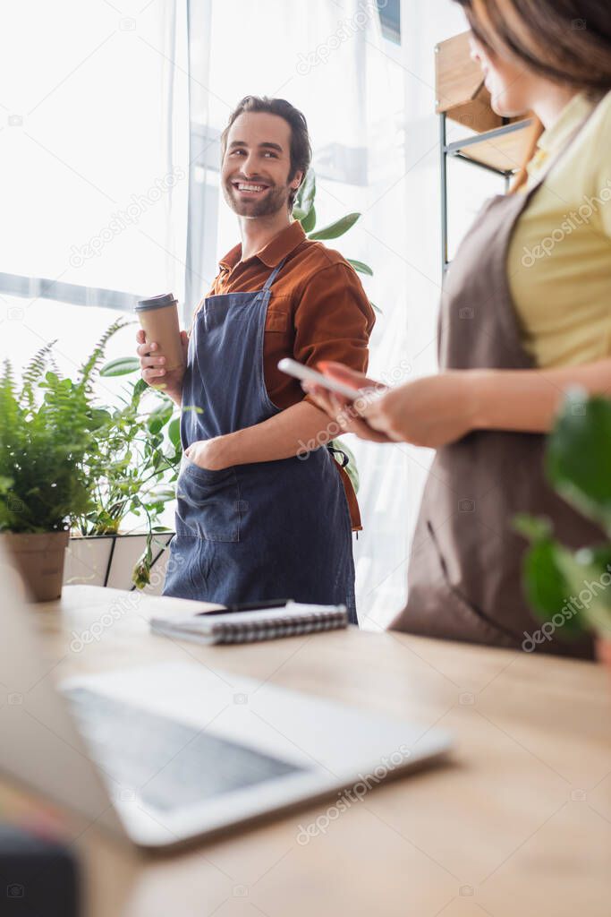 Smiling retailer with paper cup looking at colleague with smartphone in flower shop 