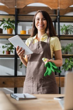 Çiçekçide gülümseyen çiçekçi eldiven ve bilgisayarın yanında dijital tablet tutuyor. 