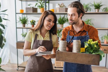 Çiçekçi gülümsüyor. İş arkadaşının yanındaki defterde çiçek saksısı ve çiçek saksısı var. 